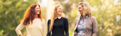 Drei Baufinanzierungsberaterinnen stehen nebeneinander im Grünen und lächeln sich einander an